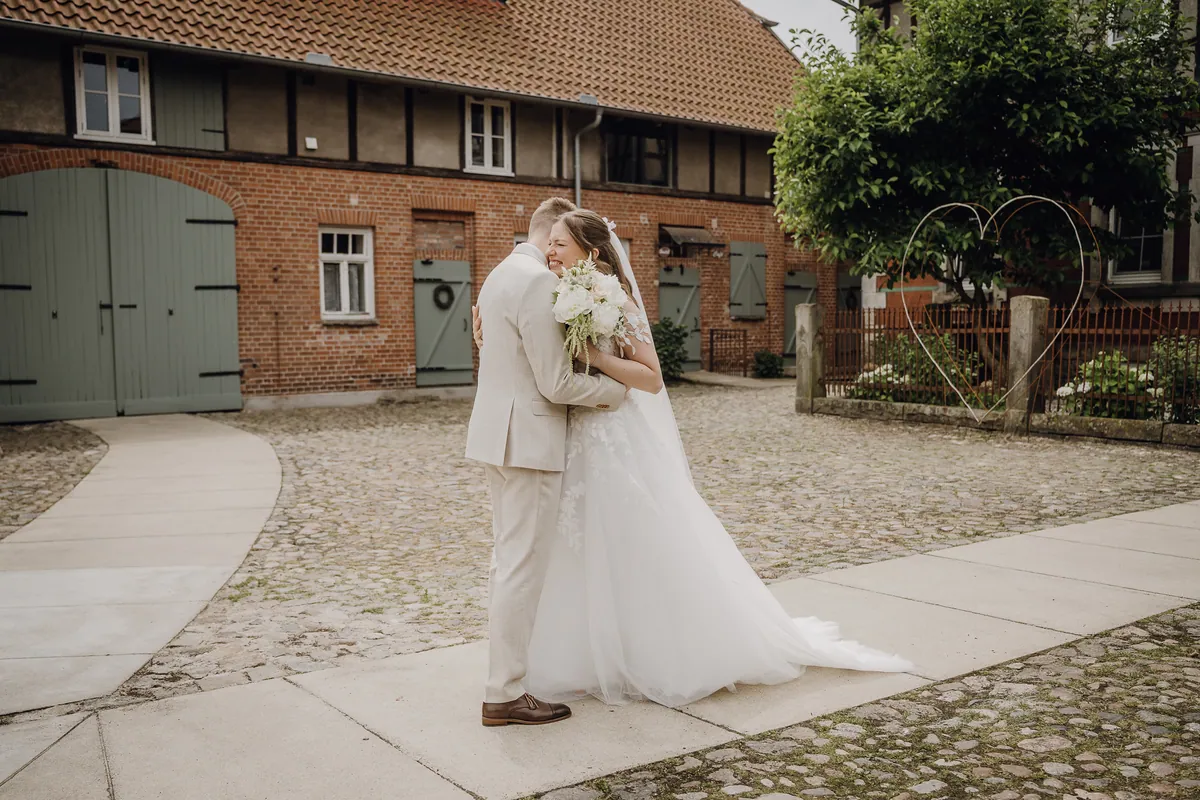 Sara lacht mit geschlossenen Augen, als Philipp sie beim First Look umarmt – auf dem gepflasterten Hof vor einem Backsteinhaus mit salbeigrünen Toren.