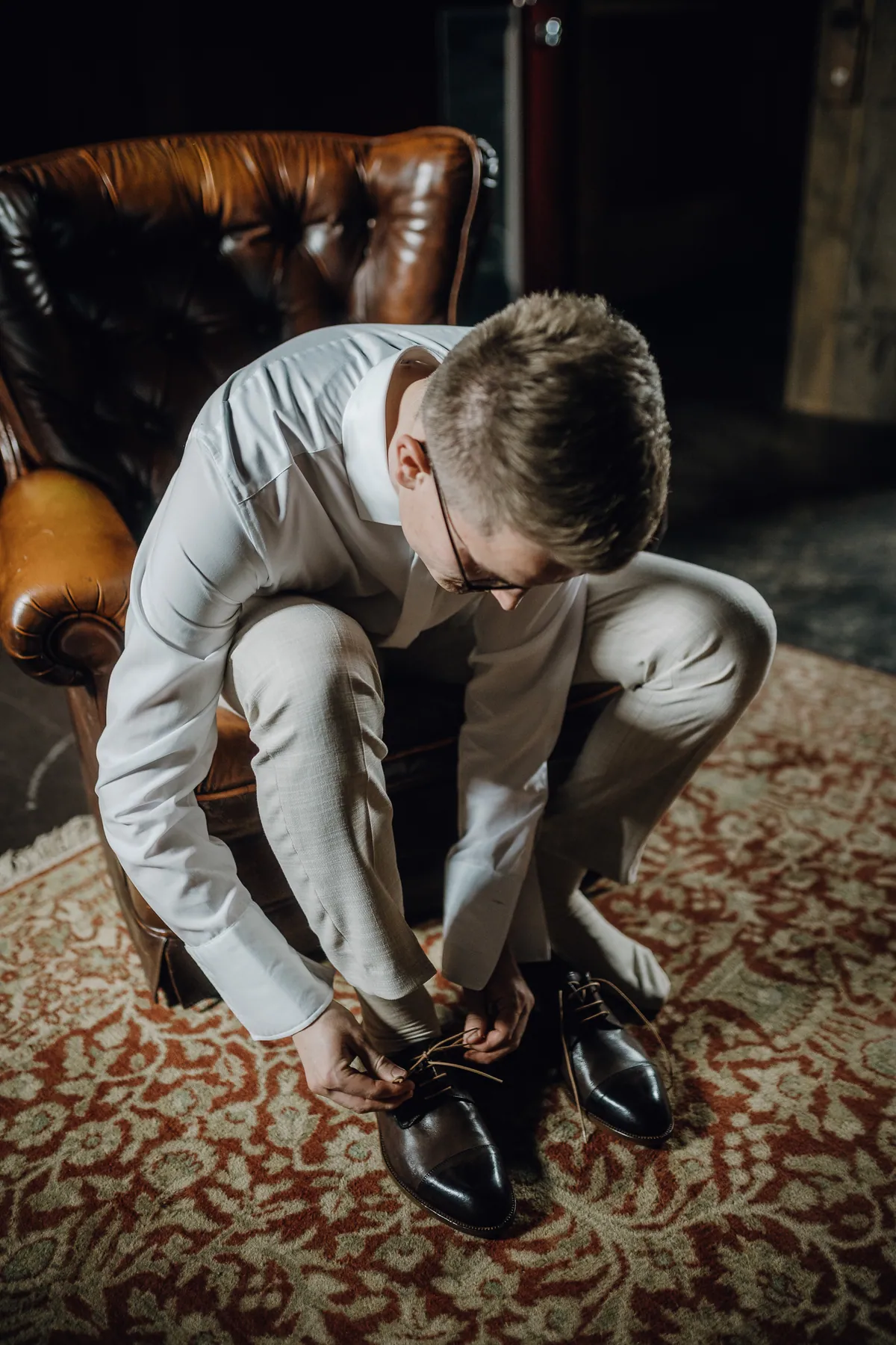Philipp sitzt im weißen Hemd und heller Anzughose auf einem braunen Ledersessel und bindet seine Lederschuhe auf einem Orientteppich.