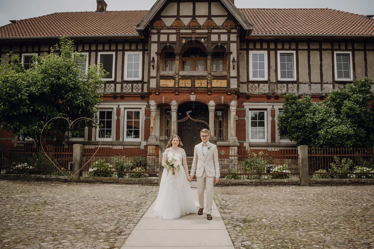 Sara und Philipp gehen Hand in Hand auf dem Weg vor einem prächtigen Fachwerkhaus mit Reliefs und Säulenportal auf die Kamera zu.