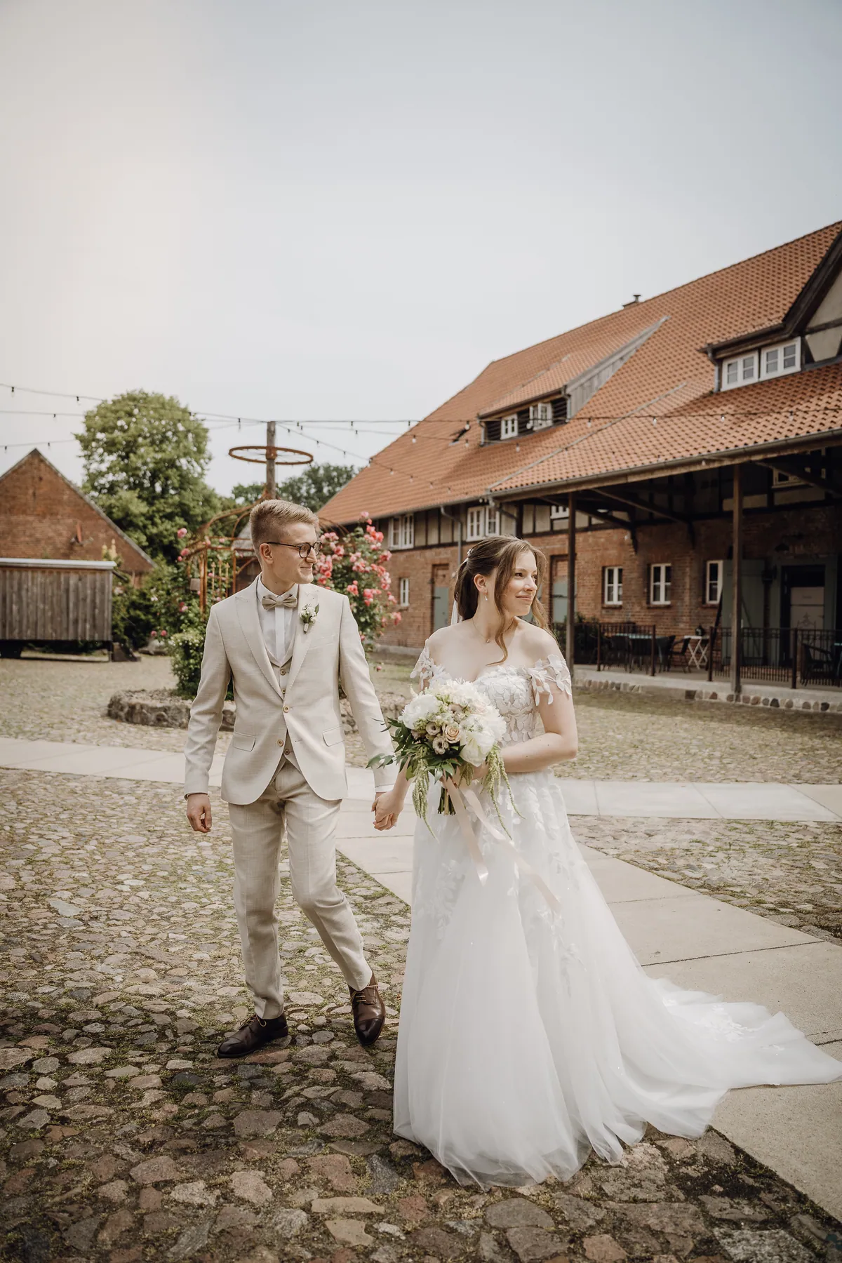 Hand in Hand gehen Braut und Bräutigam über den Hof, sie blickt lächelnd zur Seite, hinten ein Rosenpavillon und ein Backsteinhaus mit Lichterketten.