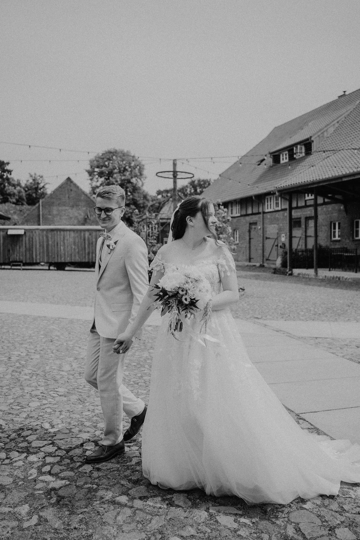 Schwarzweißaufnahme: Sara und Philipp gehen Hand in Hand über einen gepflasterten Hof, sie blickt zur Seite, über ihnen hängt eine Lichterkette vor einer Fachwerkscheune.