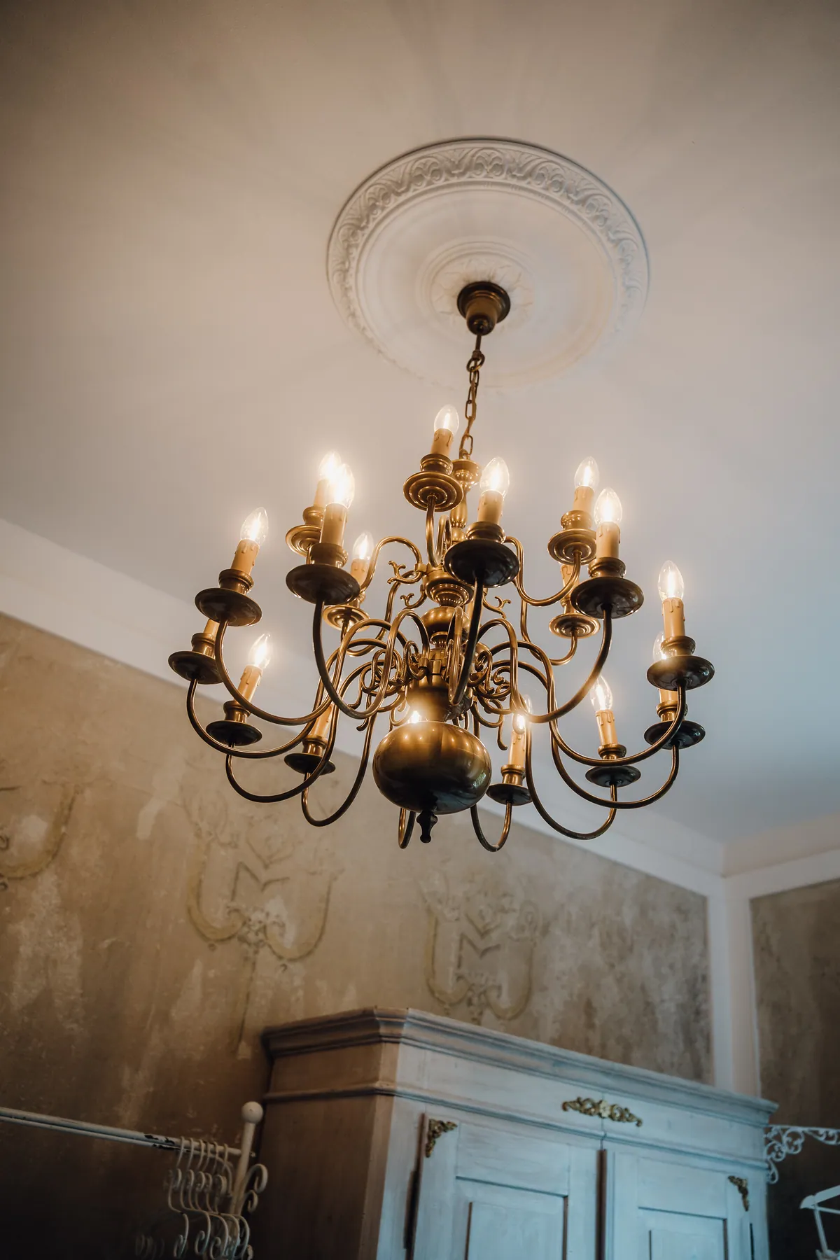 Ein Messing-Kronleuchter mit leuchtenden Kerzenlampen hängt unter einer Stuckrosette, darunter ein weißer Bauernschrank vor verzierten Patina-Wänden.