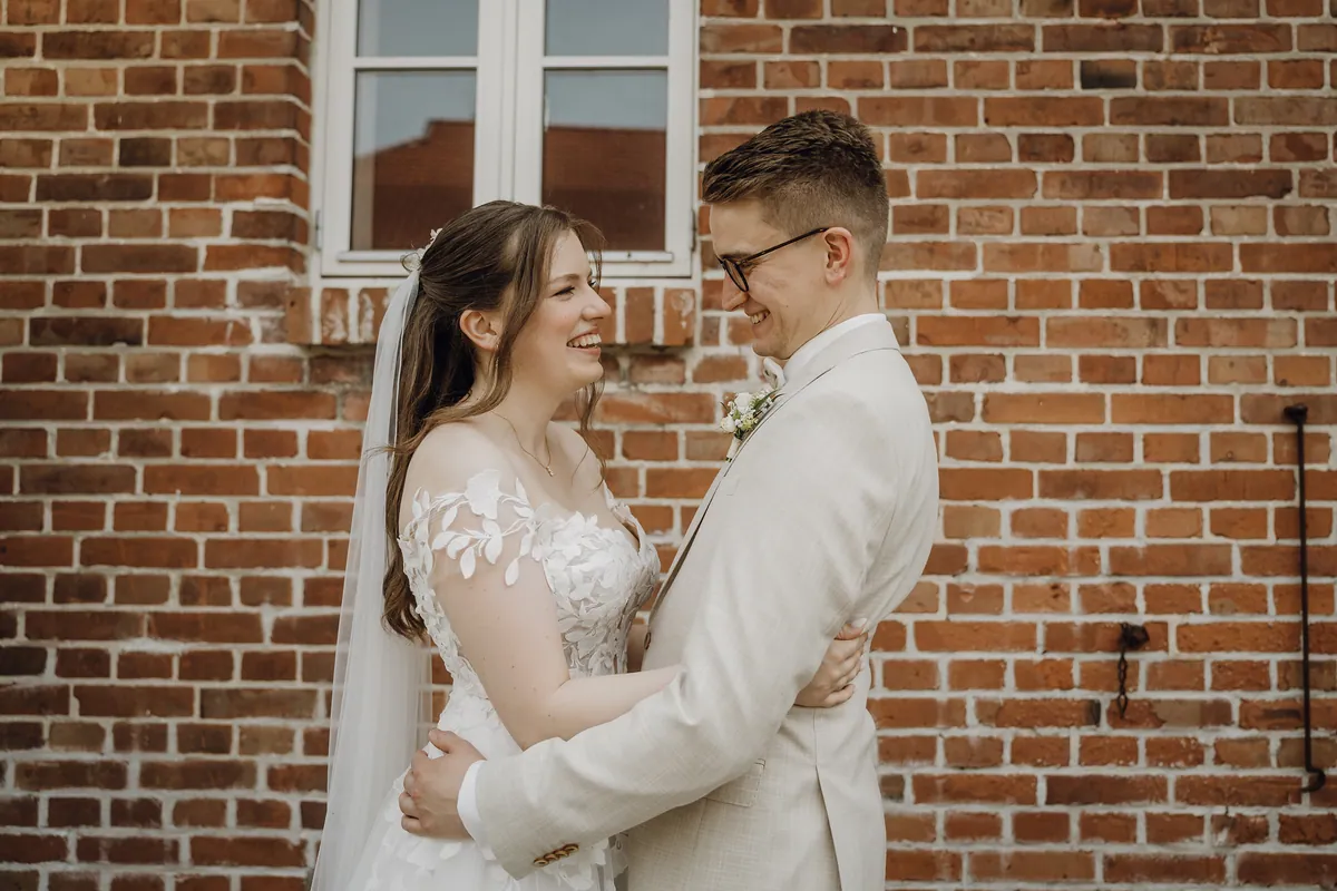 Sara und Philipp halten sich vor einer roten Backsteinwand unter einem weißen Sprossenfenster im Arm und lachen sich an, sie trägt Schleier und halboffenes Haar.