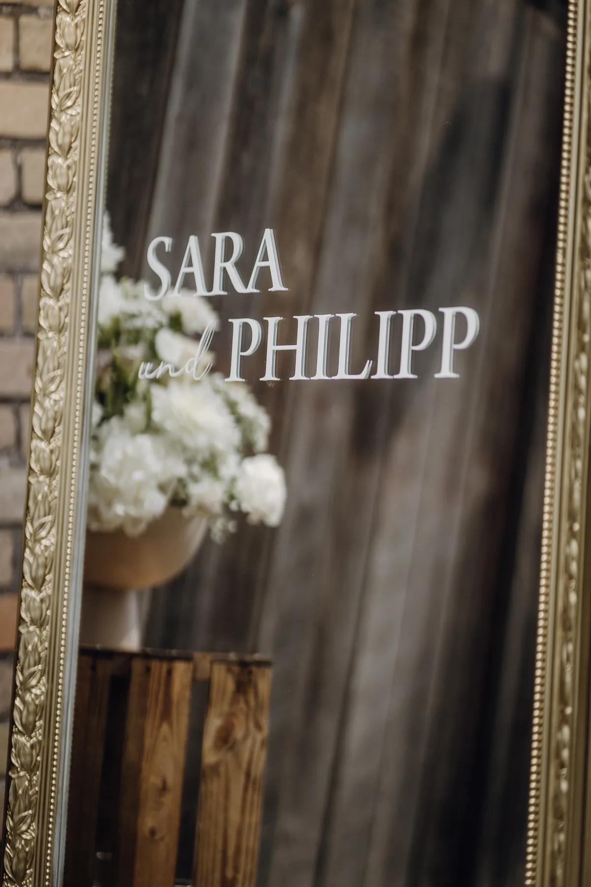 Ein Spiegel im verzierten Goldrahmen trägt den weißen Schriftzug «Sara und Philipp», im Spiegelbild sind weiße Blumen in einer Schale auf einer Holzkiste zu sehen.
