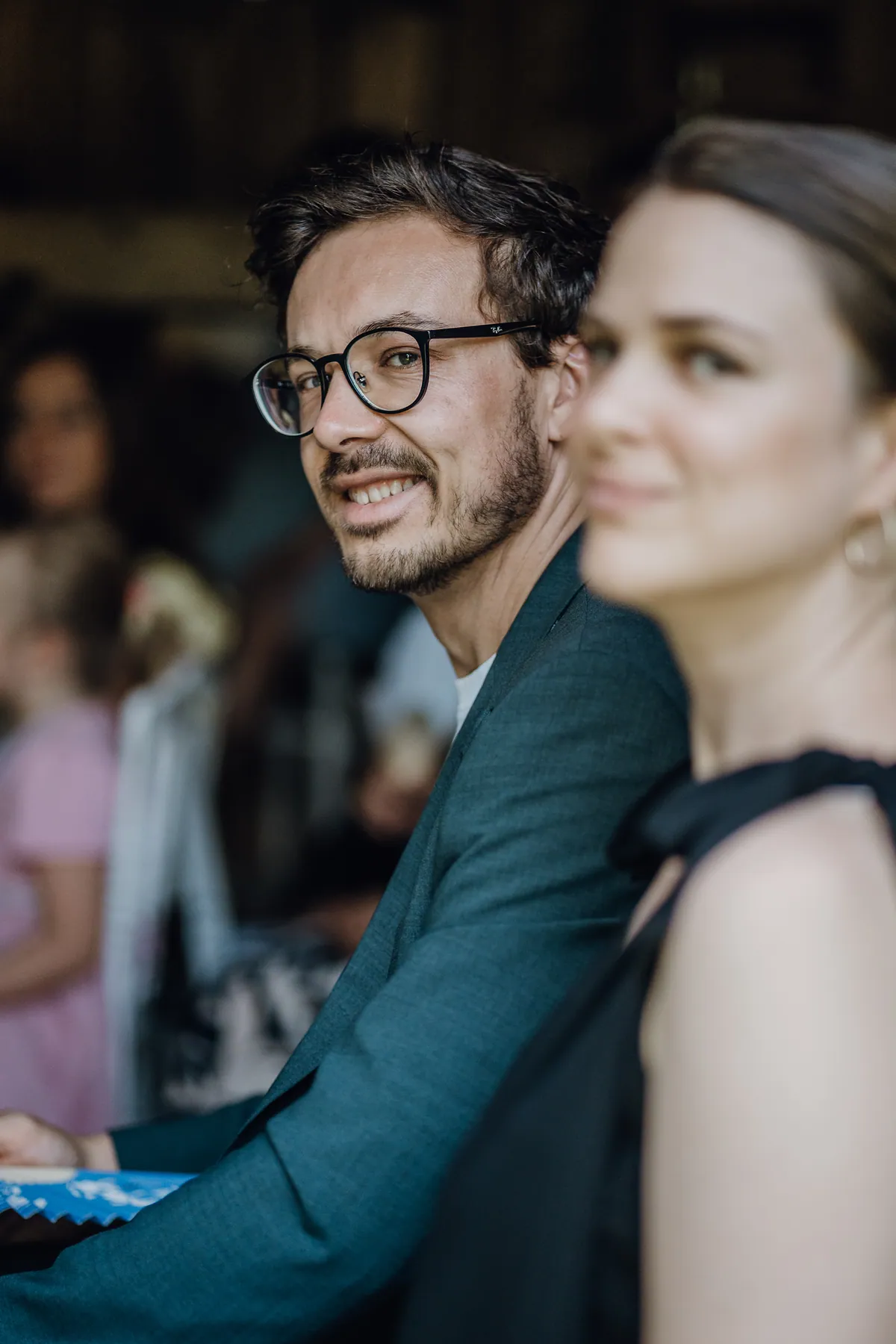 Ein Gast mit Brille und Bart im petrolfarbenen Sakko lächelt über die Schulter in die Kamera, im Vordergrund unscharf eine Frau im schwarzen Kleid.
