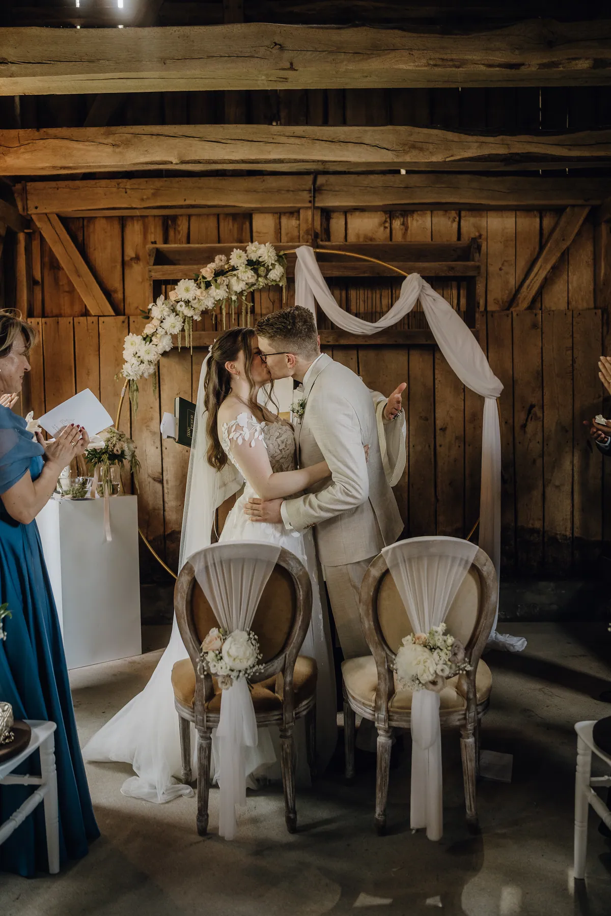 Nach dem Jawort küssen sich Sara und Philipp vor einem goldenen Ringbogen mit weißen Blumen in der Holzscheune, er reckt jubelnd einen Arm hoch, links applaudiert eine Frau im blauen Kleid.