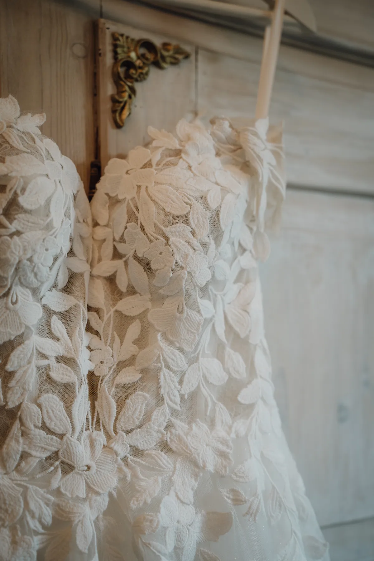 Detail des Brautkleids: florale Spitzenapplikationen auf zartem Tüll am herzförmigen Ausschnitt, dahinter eine Schranktür mit goldenem Beschlag.