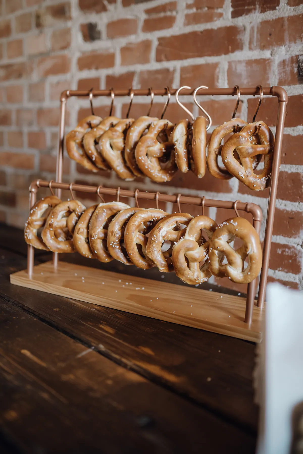 Laugenbrezeln hängen an Kupferhaken in einem zweistöckigen Ständer aus Kupferrohr, der auf einem dunklen Holztisch vor einer Backsteinwand steht.