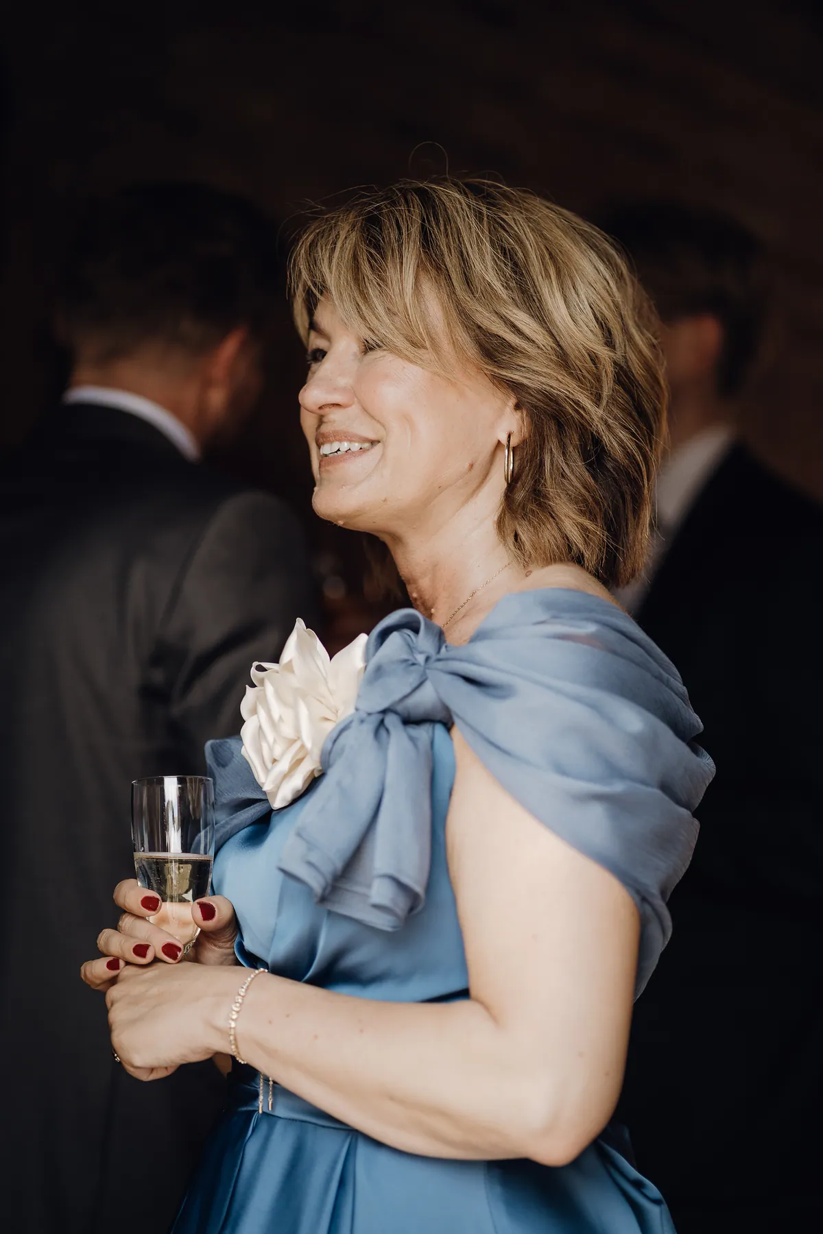 Ein Gast im hellblauen Satinkleid mit Schulterschleife und weißer Stoffblume lacht im Profil und hält ein Glas Sekt mit beiden Händen.