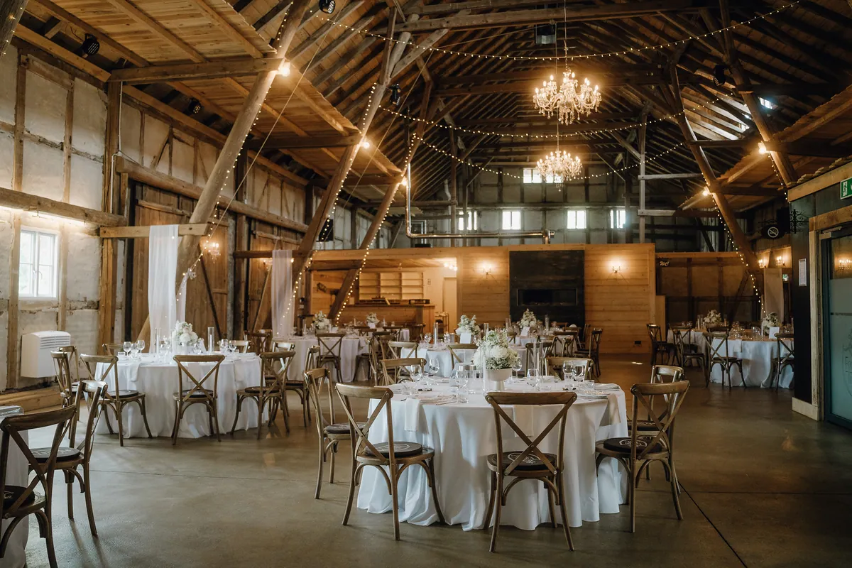 Festsaal in einer hohen Holzscheune mit runden, weiß eingedeckten Tischen, Holzstühlen mit Kreuzlehne, Kristalllüstern und Lichterketten unter dem Dachgebälk.