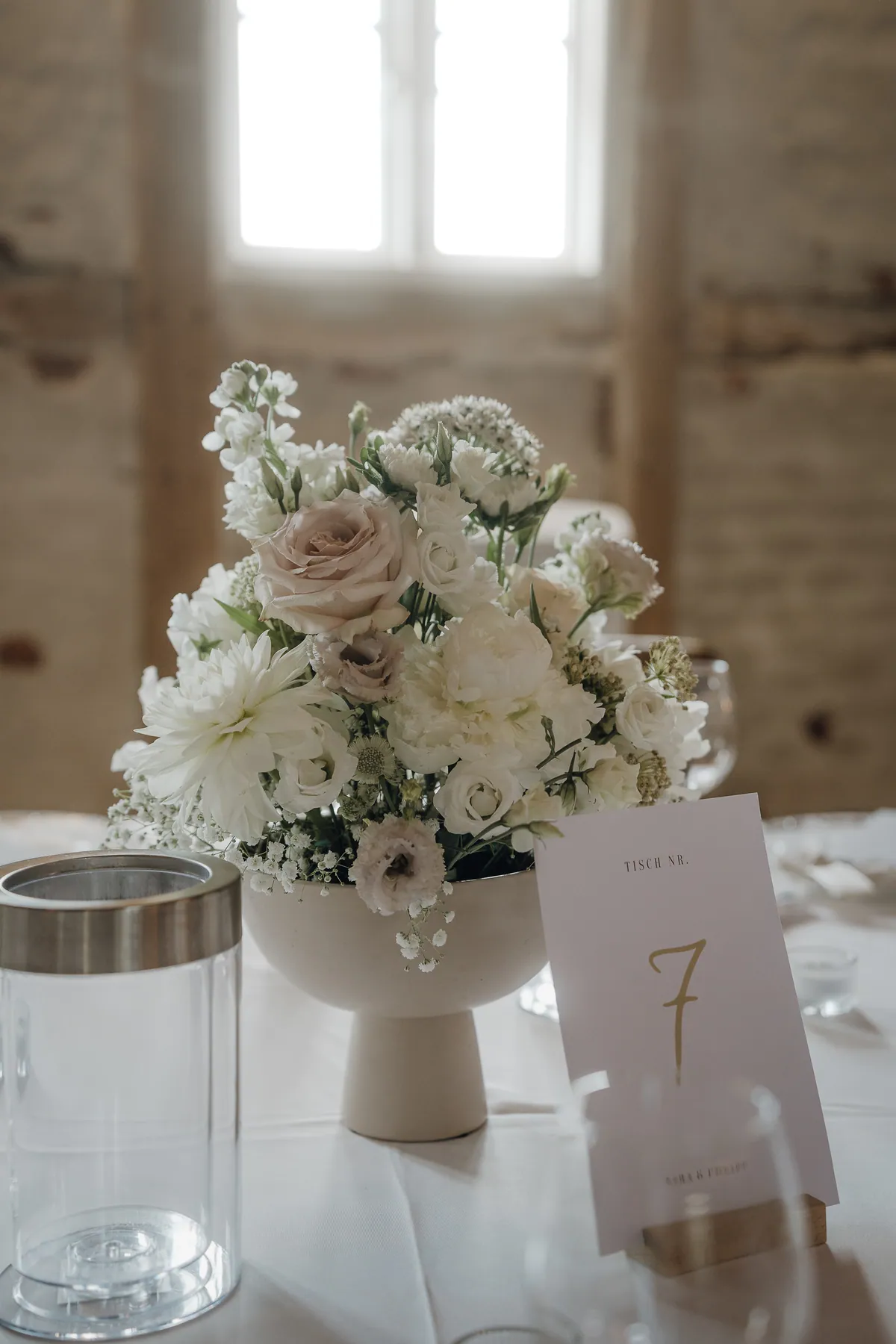 Tischdeko in Weiß und Altrosa: Rosen, eine Dahlie und Schleierkraut in einer Keramikschale auf Fuß, daneben eine Karte mit goldener Tischnummer 7 und ein Windlicht.