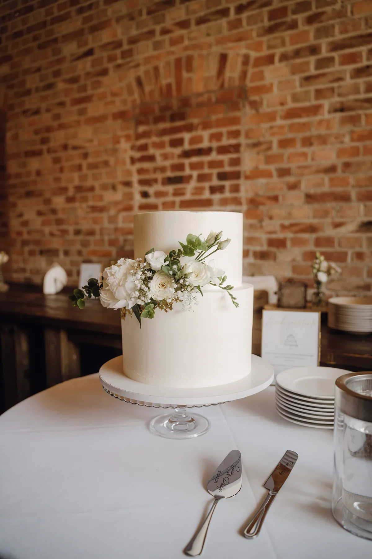 Eine zweistöckige weiße Hochzeitstorte mit Pfingstrose, Rosen und Schleierkraut steht auf einer Glasetagere vor einer Backsteinwand, davor ein gravierter Tortenheber.