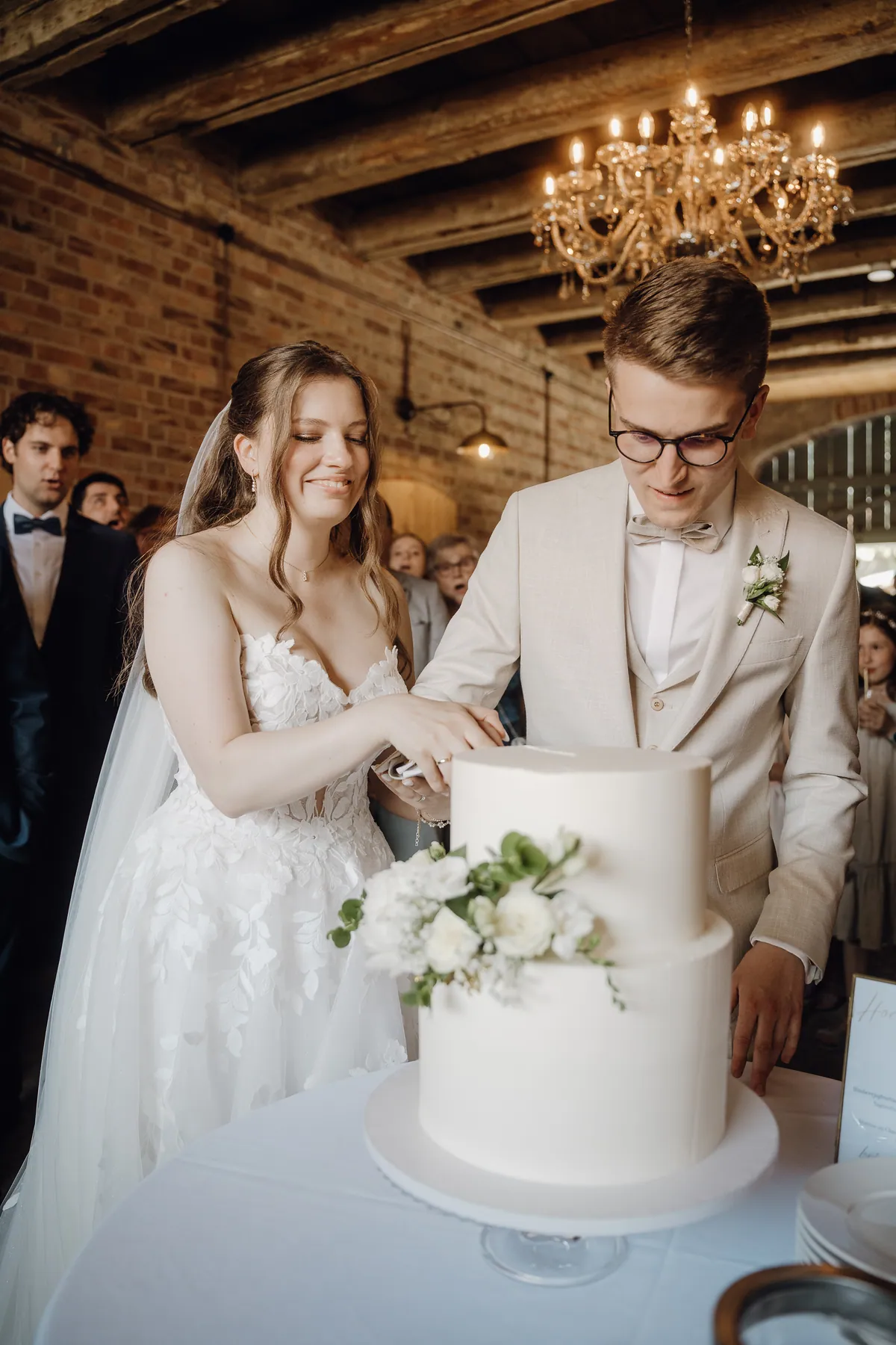 Sara und Philipp schneiden gemeinsam die weiße Hochzeitstorte an, über ihnen hängt ein Kristallleuchter unter Holzbalken, hinten schauen Gäste zu.