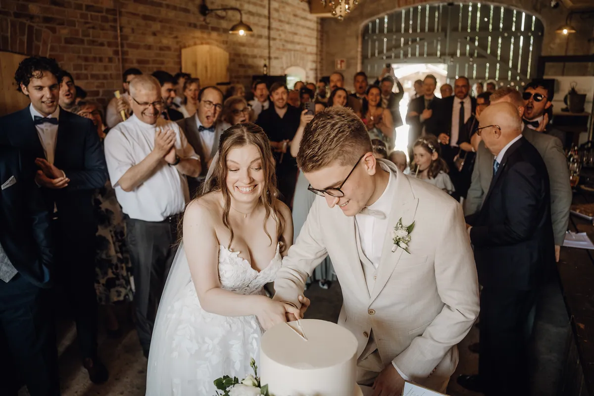 Unter dem Jubel der Gäste schneidet das Brautpaar lachend die Torte an, hinter ihnen klatschen und filmen die Umstehenden vor einem offenen Scheunentor.