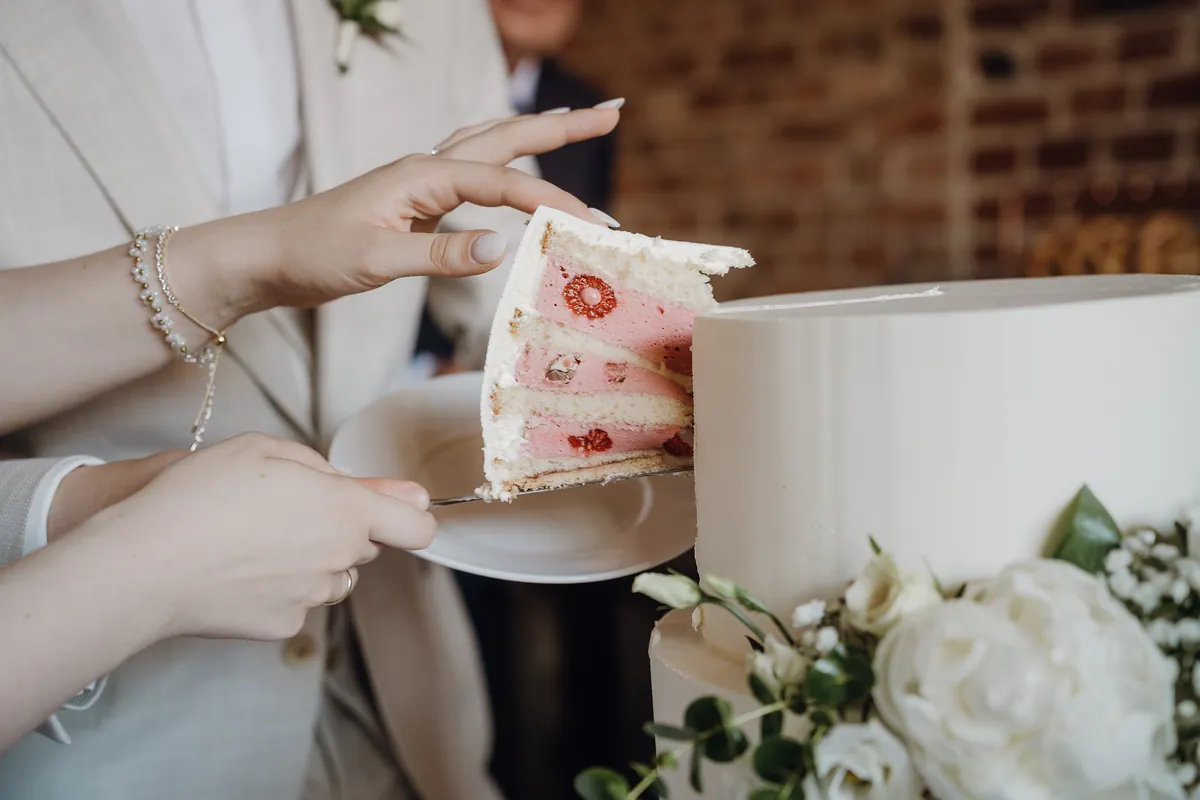 Nahaufnahme: Zwei Hände heben das erste Tortenstück auf einen Teller, im Anschnitt zeigen sich helle Biskuitböden und rosa Creme mit ganzen Himbeeren.