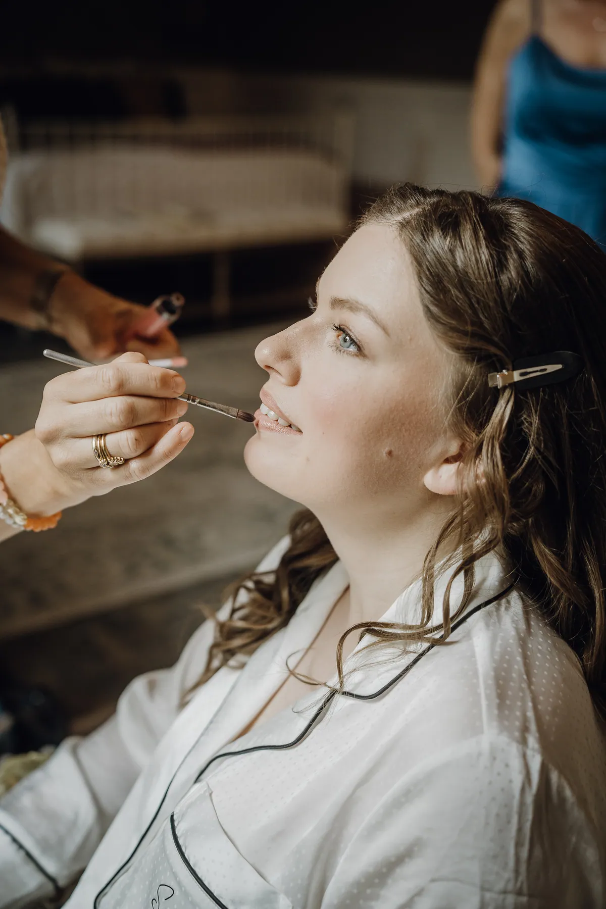 Die Braut sitzt im weißen Pyjama mit gewelltem Haar und Haarklammer, eine Visagistin trägt ihr mit einem feinen Pinsel Lippenfarbe auf.