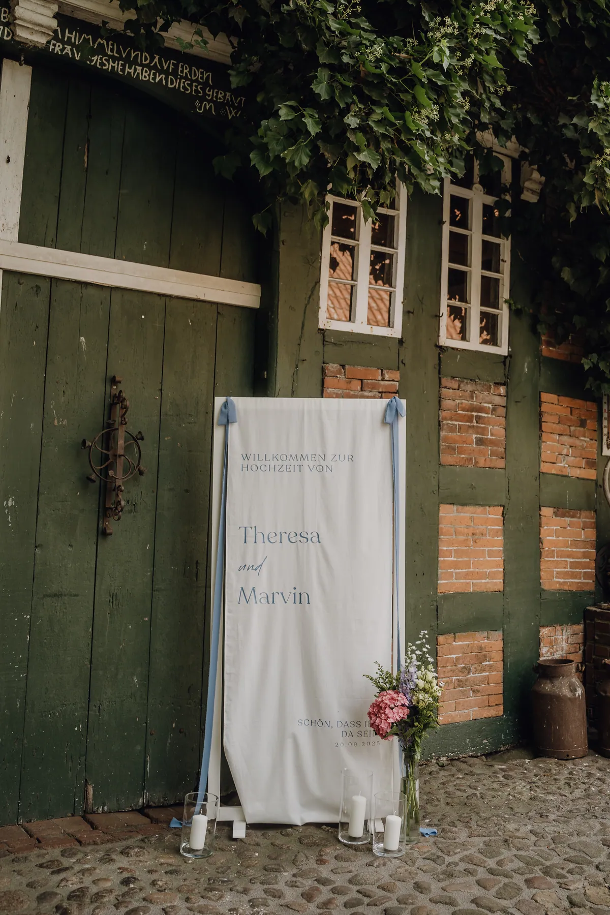 Ein Stoffbanner mit «Willkommen zur Hochzeit von Theresa und Marvin» lehnt an einem grünen Fachwerktor, davor Windlichter und ein kleiner Blumenstrauß.