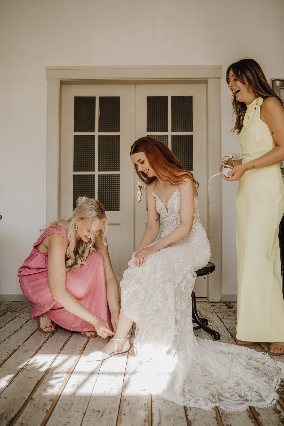 Eine Freundin in Rosa hockt vor der lächelnden Braut und schließt ihr den Schuh, eine zweite Freundin im gelben Kleid steht lachend daneben.