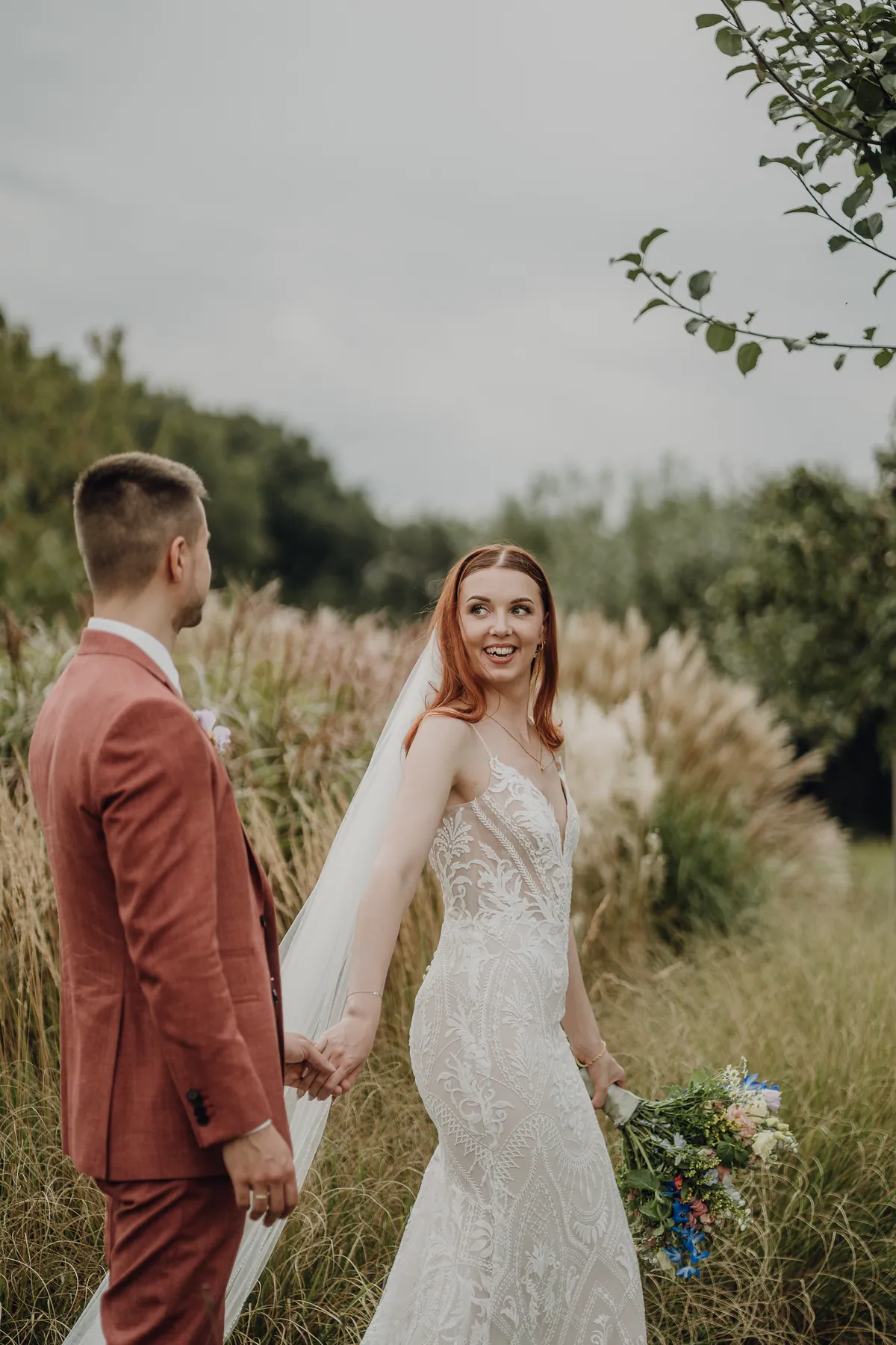 Hand in Hand mit Marvin dreht sich Theresa im Spitzenkleid lachend zu ihm um, hinter ihr stehen hohe Ziergräser mit hellen Rispen.