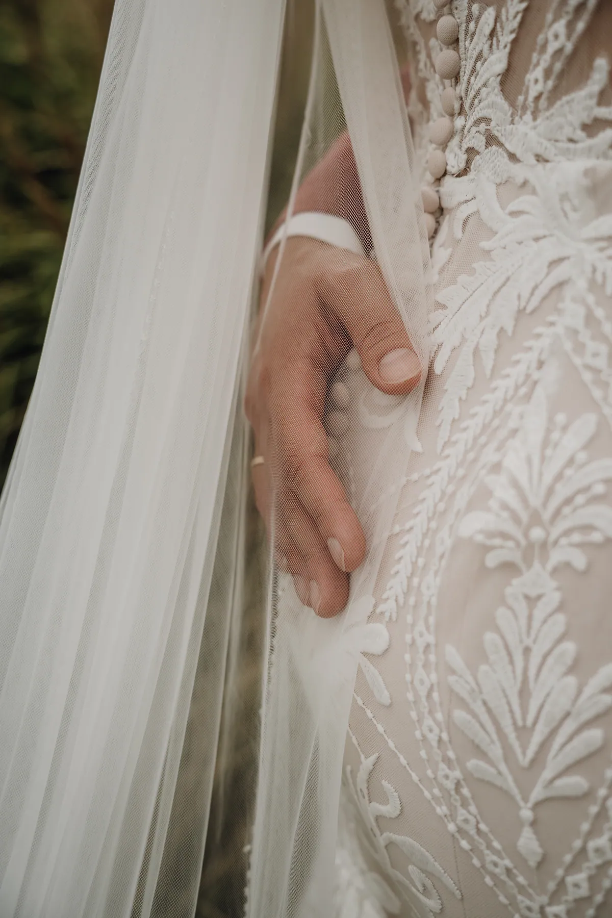 Detail: Durch den feinen Tüllschleier ist die Hand des Bräutigams mit goldenem Ehering zu sehen, sie liegt auf der Spitze und den Stoffknöpfen des Kleids.