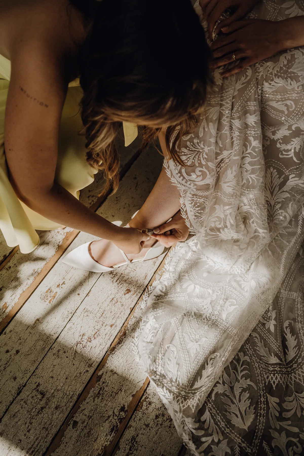 Von oben: Eine Freundin im gelben Kleid schließt das Riemchen am weißen Brautschuh, das Spitzenkleid fällt in Licht und Schatten über die Dielen.
