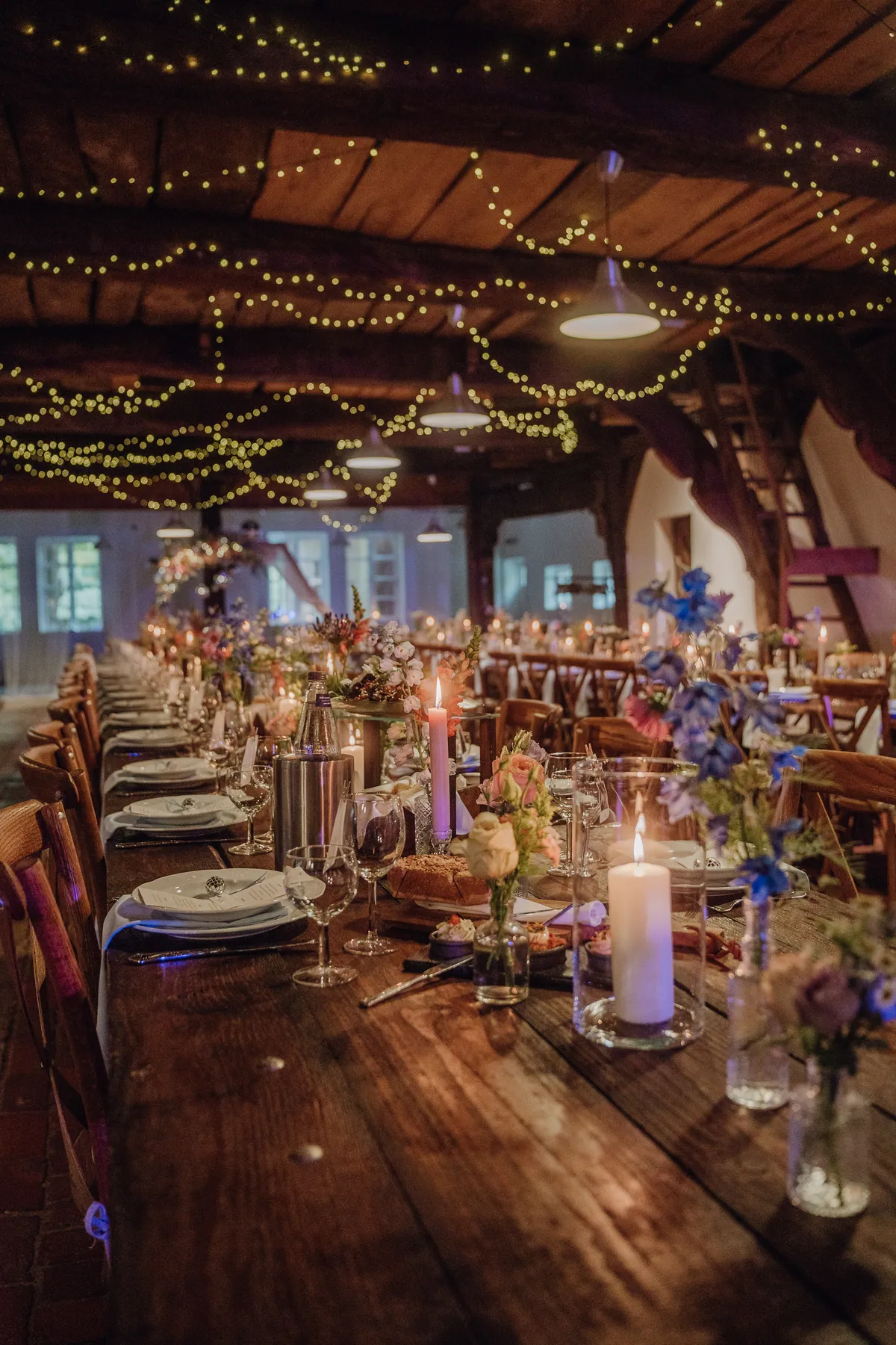 Lange Holztafel unter Lichterketten und dunklen Deckenbalken, gedeckt mit weißen Tellern, brennenden Kerzen und Wiesenblumen in kleinen Glasvasen.