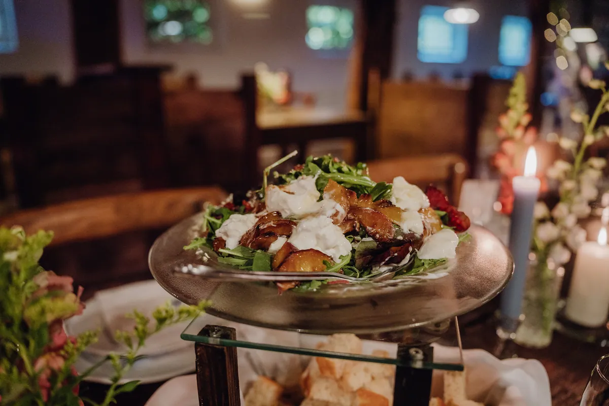 Auf einer Glasetagere steht ein Salat mit Rucola, Burrata und gebratenen Fruchtspalten, darunter ein Korb mit Brot, rechts brennen Kerzen.