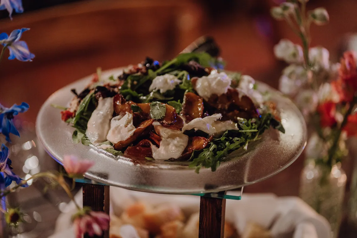 Nahaufnahme des Salats auf der Glasplatte: Rucola mit cremigem Käse und glasierten Fruchtspalten, am Rand unscharf blaue und rosa Blüten.
