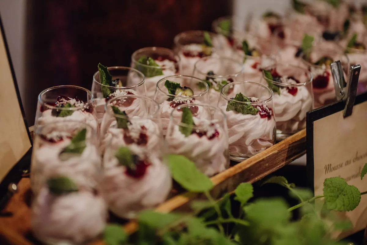 Dessert in kleinen Gläsern: rosa Mousse mit roter Fruchtsoße, Kokosraspeln und Minzblatt, aufgereiht auf einem Holztablett neben einem Schild.
