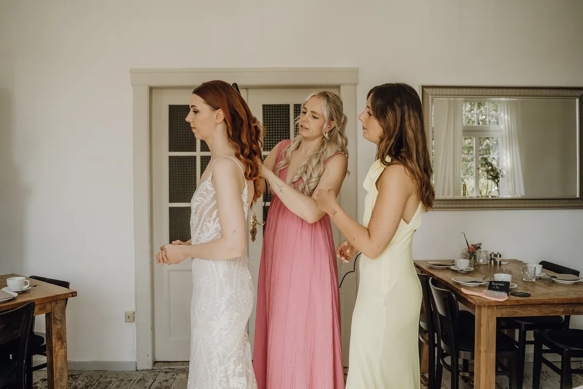 Die Braut steht im Spitzenkleid im hellen Raum, die Freundin in Rosa legt ihr mit dem Lockenstab die Haare, die Freundin in Gelb schaut zu.