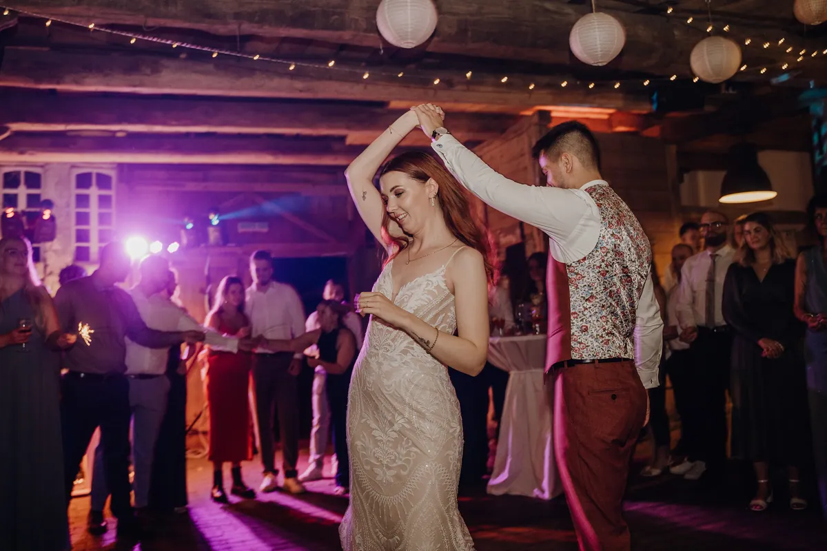 Marvin, dessen Weste ein geblümtes Rückenteil hat, dreht Theresa unter seinem erhobenen Arm, sie lächelt mit geschlossenen Augen, im Hintergrund leuchtet buntes Partylicht.