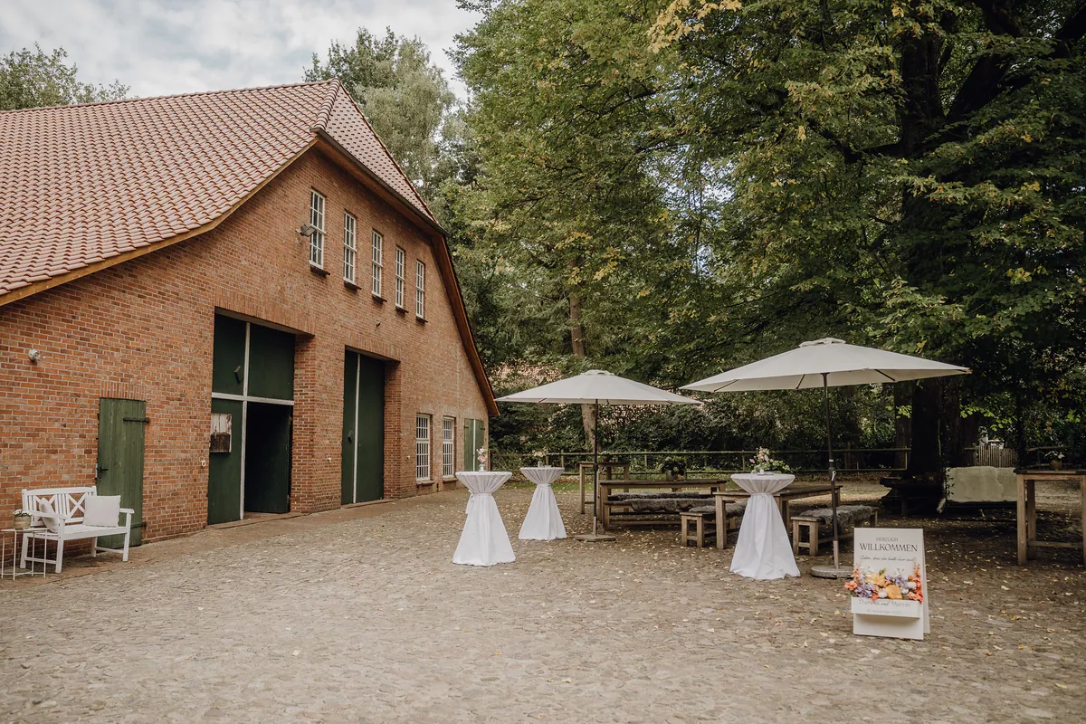 Der Innenhof von Hof Schweers am Hochzeitstag: rote Backsteinscheune mit grünen Toren, weiße Stehtische und Sonnenschirme unter alten Bäumen.