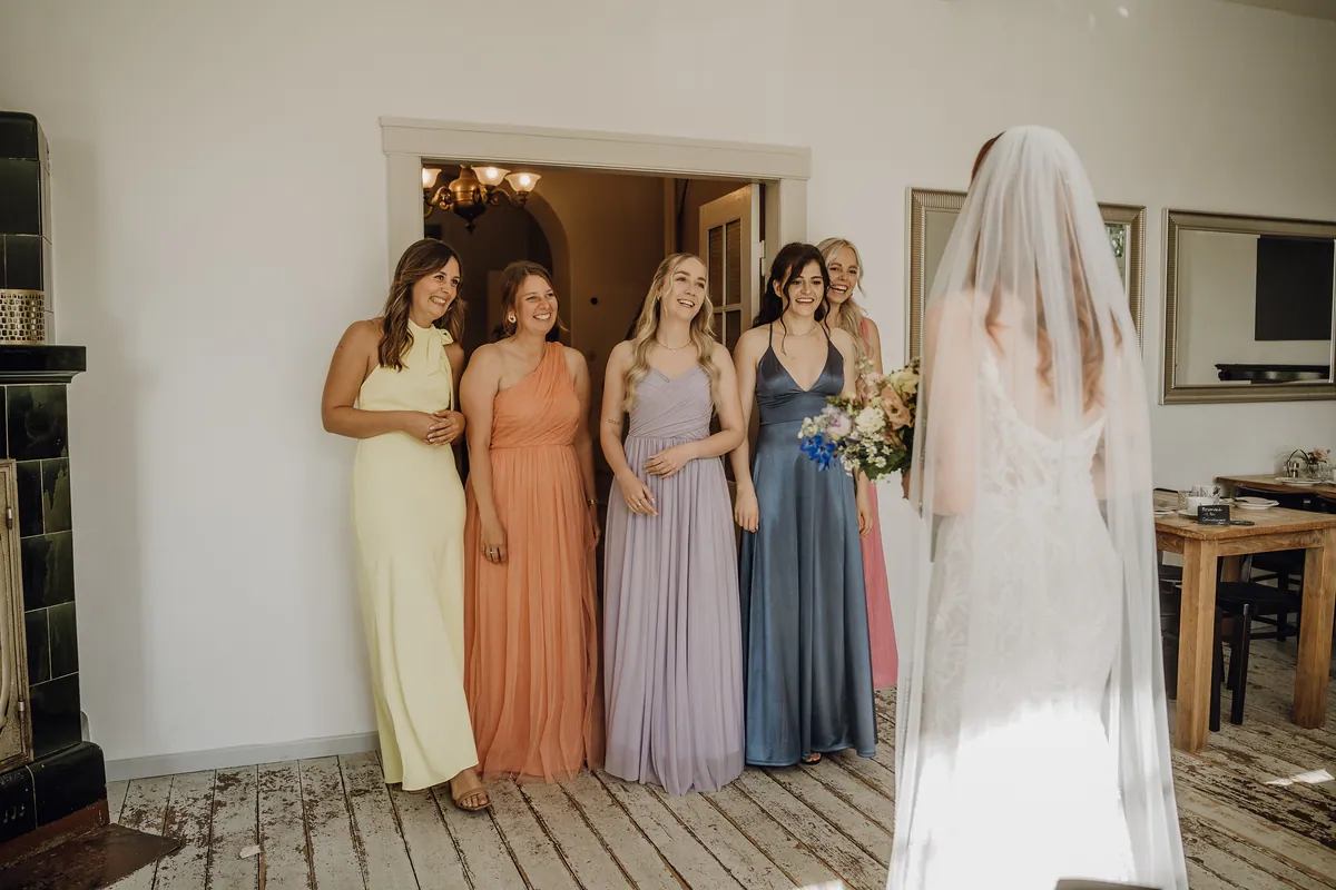 Fünf Freundinnen in Gelb, Apricot, Flieder, Blau und Rosa stehen strahlend an der Tür und schauen auf die Braut, die mit Schleier von hinten zu sehen ist.