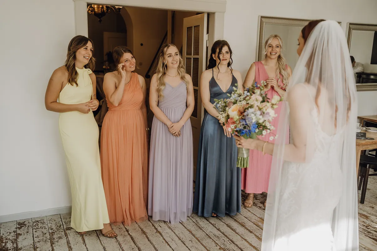 Die Freundinnen in pastellfarbenen Kleidern sehen die Braut mit Strauß und Schleier an, eine im apricotfarbenen Kleid wischt sich eine Träne weg.