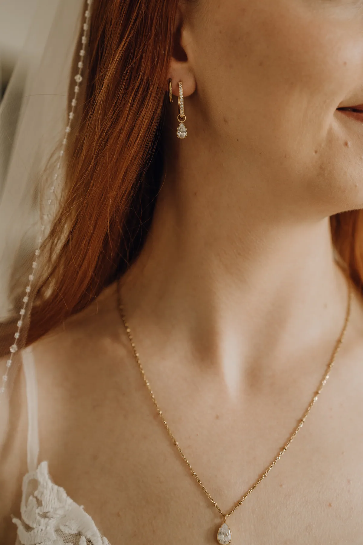 Nahaufnahme: Am Ohr der Braut hängen goldene Creolen mit Tropfenanhänger, dazu eine feine Goldkette, daneben fällt das rote Haar unter dem Perlenschleier.