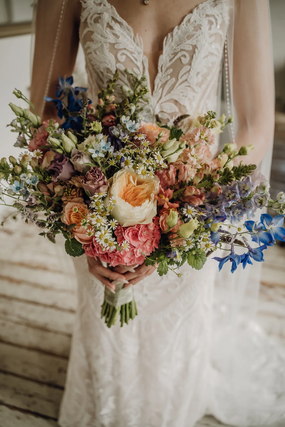 Die Braut hält ihren Strauß vor dem Spitzenkleid: Pfirsichrosen, korallenrosa Blüten, Kamille und blauer Rittersporn, über die Schultern fällt der Schleier.