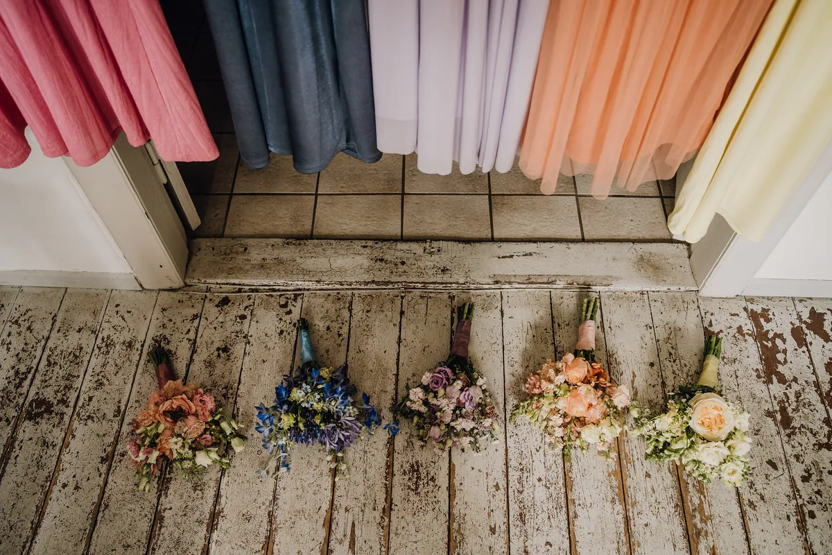Von oben: Fünf kleine Sträuße in Rosa, Blau, Flieder, Apricot und Creme liegen auf abgeblätterten Dielen, darüber hängen Kleider in denselben Farben.