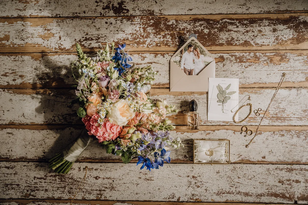Auf verwittertem Holz liegen der bunte Brautstrauß, eine Einladung mit Paarfoto im Umschlag, ein Parfümflakon, Goldschmuck und die gläserne Ringbox.