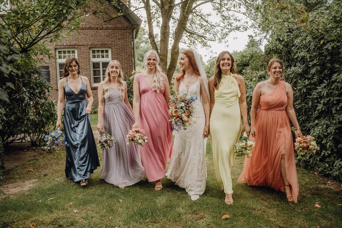 Im Gehen schaut die Braut lächelnd zu ihrer Freundin im rosa Kleid, rundherum die Brautjungfern in Blau, Flieder, Gelb und Apricot, dahinter ein Backsteinhaus.