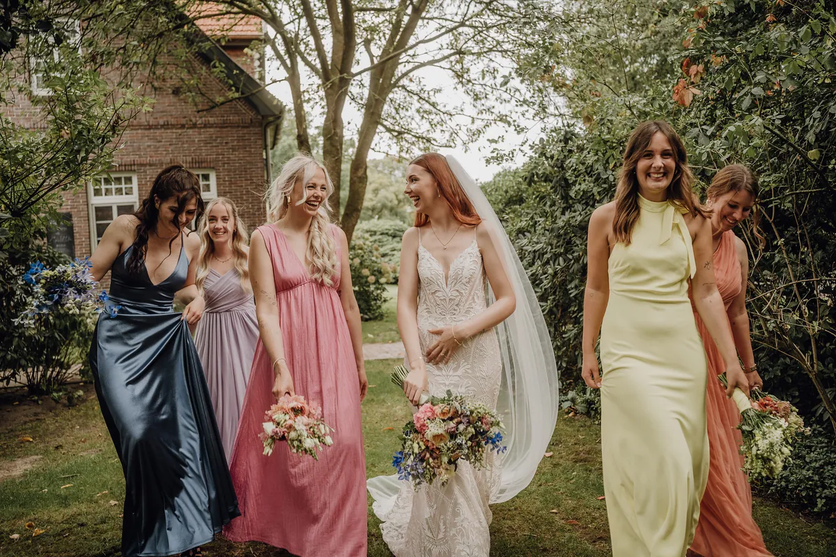 Nah dran: Die Braut und die Freundin im rosa Kleid lachen sich im Gehen an, links rafft eine Brautjungfer ihr blaues Satinkleid.
