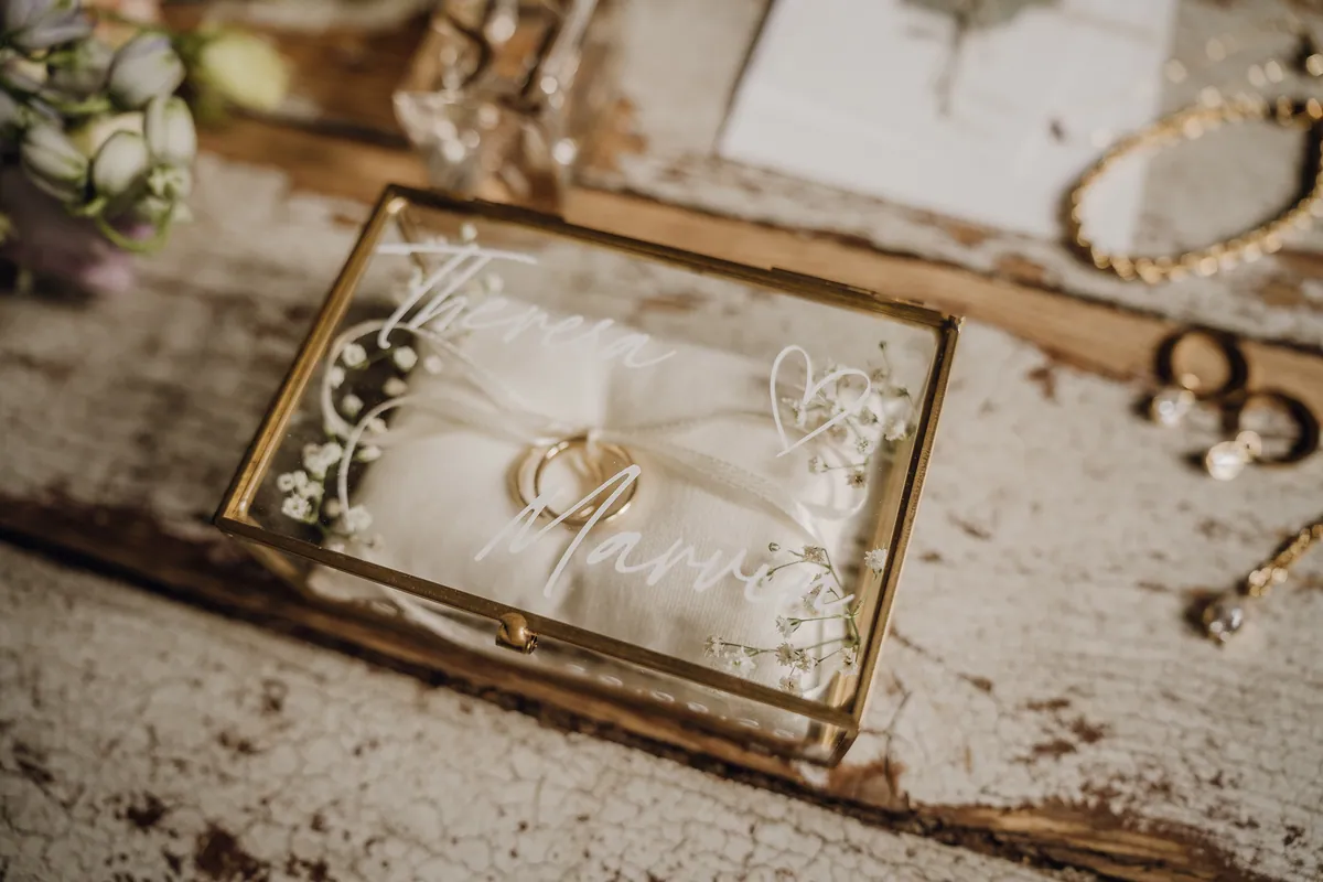 Nahaufnahme: In einer goldgerahmten Glasbox mit den weißen Schriftzügen «Theresa» und «Marvin» liegen zwei goldene Eheringe auf einem Leinenkissen.