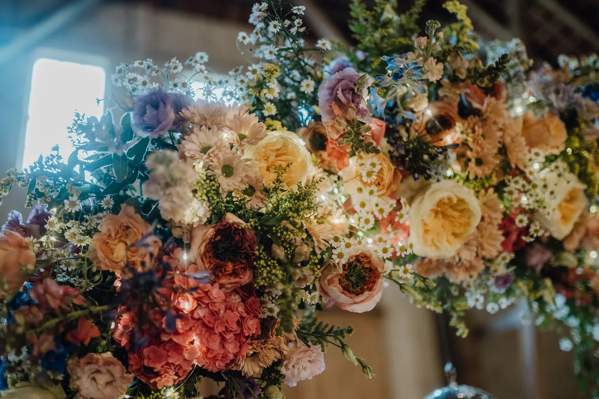 Nahaufnahme des Blumenbogens: Pfirsichfarbene Rosen, rosa Hortensien, kleine weiße Margeriten und blauer Rittersporn, dazwischen funkeln winzige Lichter.