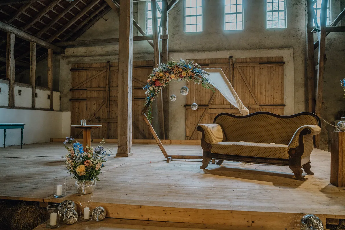 In einer Scheune steht auf der Holzbühne ein Traubogen mit bunten Blumen, Discokugeln und hellem Tuch, daneben ein altes Sofa mit senfgelbem Polster.