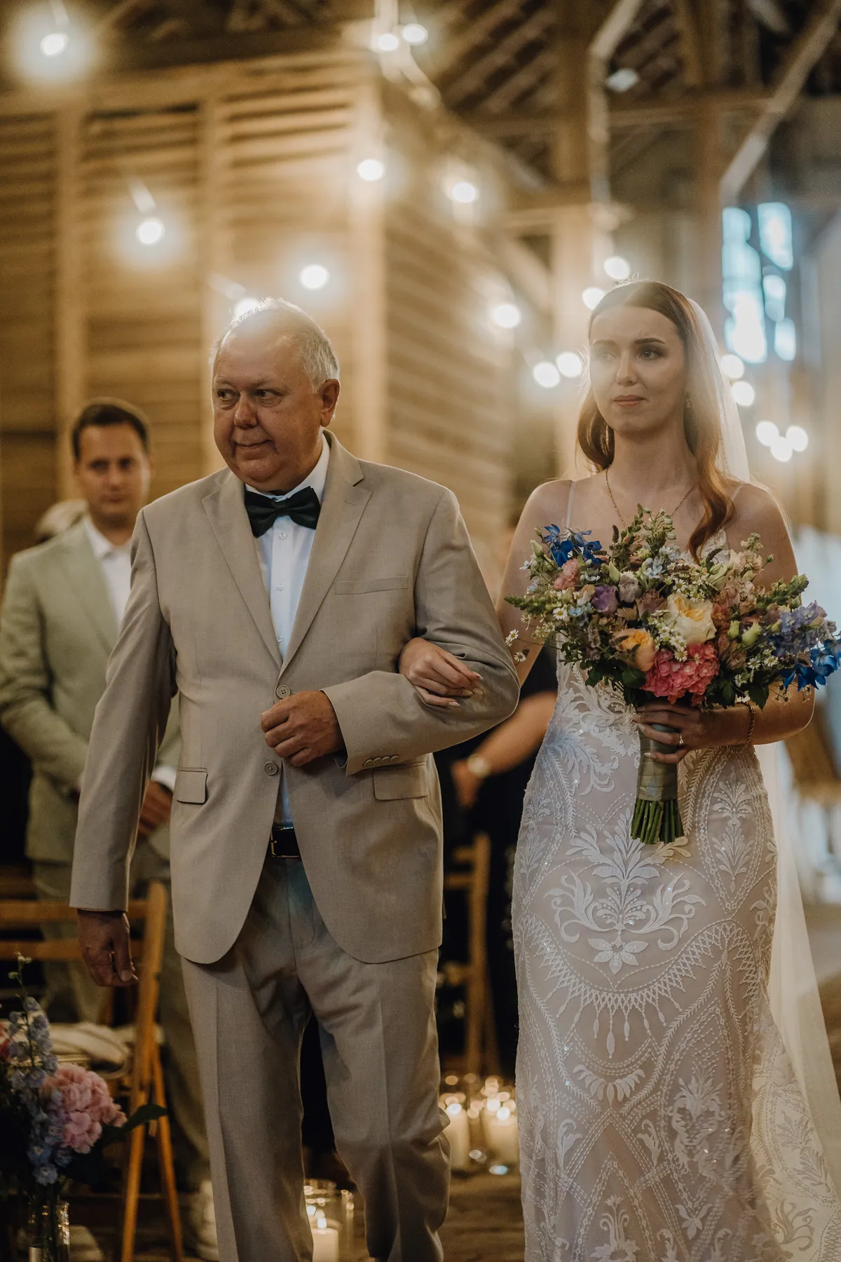 Mit bewegtem Blick geht die Braut eingehakt neben dem Herrn im beigen Anzug, ihr Strauß mit blauem Rittersporn und Hortensie leuchtet vor dem Spitzenkleid.