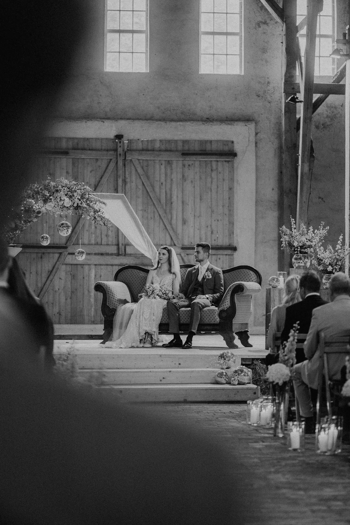Schwarzweißaufnahme: Zwischen unscharfen Silhouetten hindurch sitzt das Paar auf dem Sofa vor dem großen Holztor, rechts die Gäste am Kerzengang.