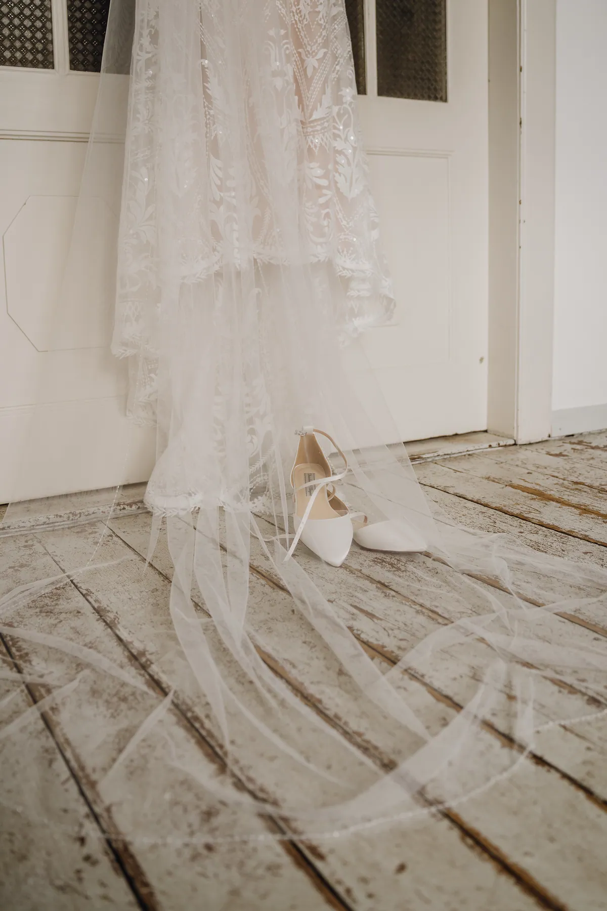 Das Brautkleid aus Spitze hängt mit langem Schleier an einer weißen Kassettentür, darunter stehen spitze weiße Brautschuhe mit Riemchen auf den Dielen.
