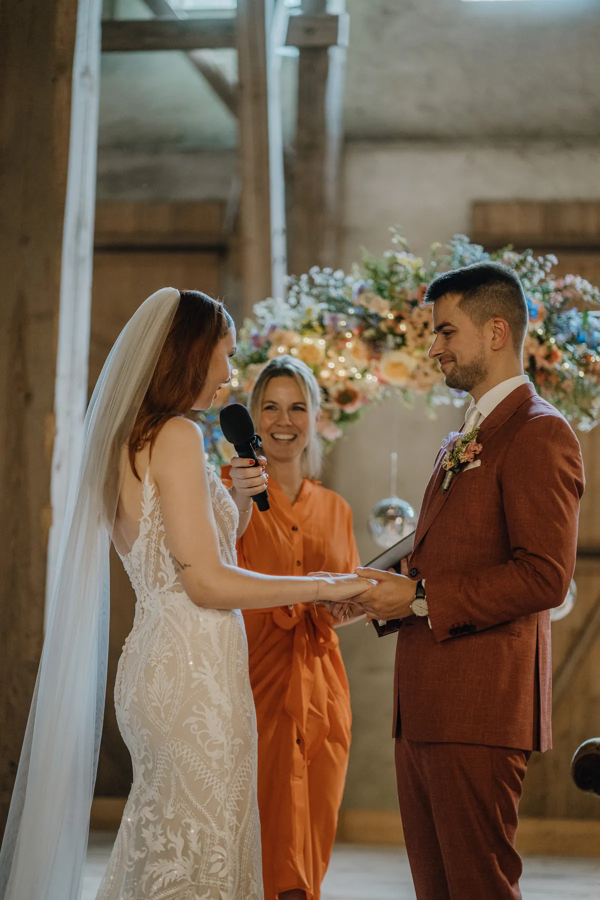 Theresa hält Marvins Hand und spricht ins Mikrofon, er lächelt sie an, zwischen beiden lacht eine Frau im orangefarbenen Kleid vor dem Blumenbogen.