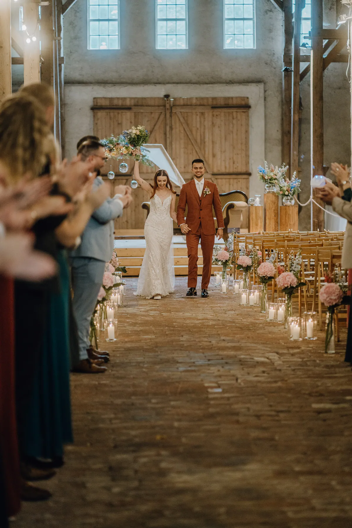 Die Braut reckt den Strauß in die Luft und geht Hand in Hand mit dem Bräutigam den Backsteingang entlang, gesäumt von Kerzen, rosa Hortensien und klatschenden Gästen.