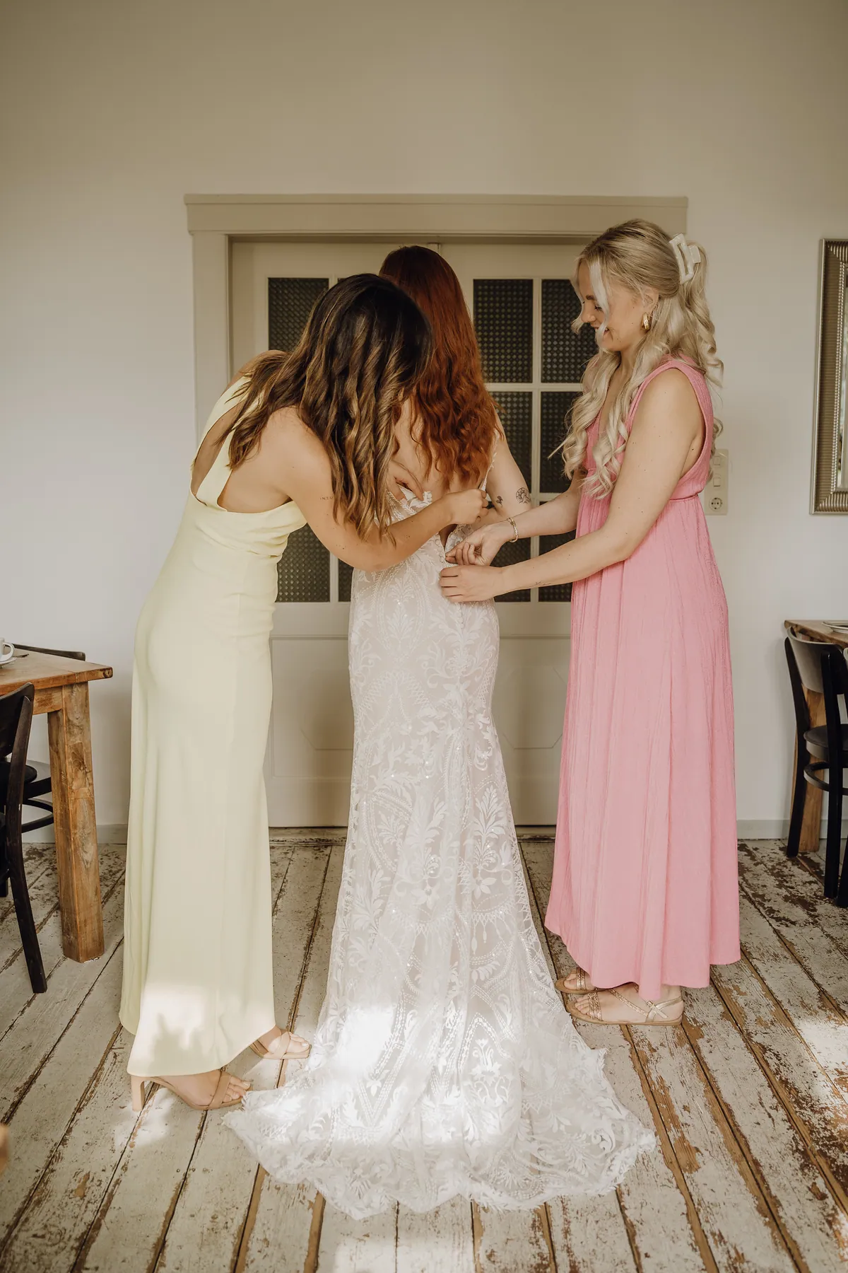 Zwei Freundinnen in gelbem und rosa Kleid schließen der rothaarigen Braut vor einer alten Sprossentür das Spitzenkleid am Rücken.