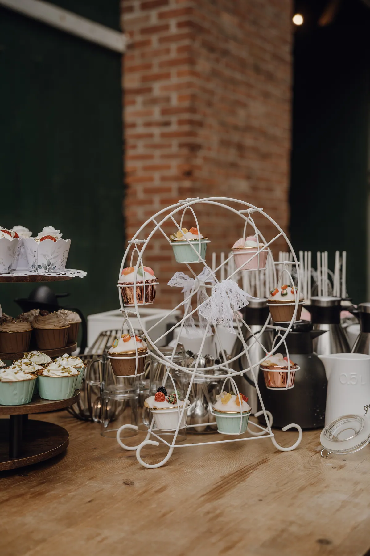 Cupcakes mit Fruchtgummis hängen in einem kleinen weißen Riesenrad aus Metall mit Spitzenschleife, dahinter Thermoskannen und ein Pfeiler aus rotem Backstein.